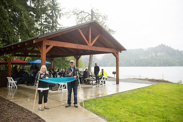 A woman and man perform a ribbon cutting ceremony with people gathered around picnic tables under an open air shelter by Lake Shannon in rainy weather.