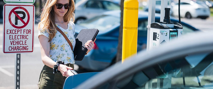 A smiling woman, wearing sunglasses, plugging a charging nozzle into her car at a workplace EV charging station