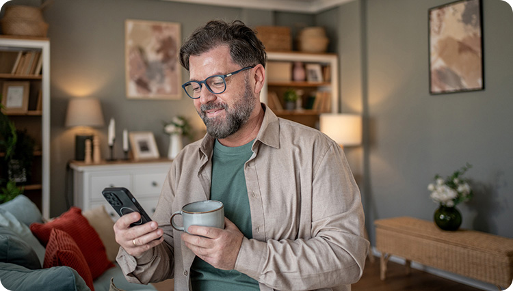 Mid adult man with glasses is standing in his living room, holding a mug and checking his phone