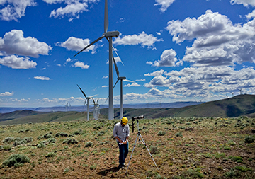 A surveyor with tripod and telescope, wearing yellow helmet, taking notes with wind turbines in background