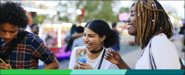A diverse trio of friends, laughing together, enjoying colorful drinks, with amusements and concession stands in the background