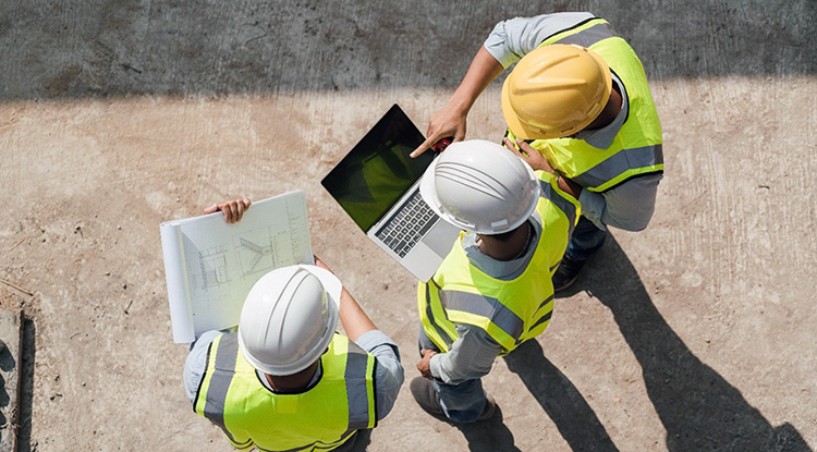 Three workers wearing hard hats and hi viz vests look at laptop screen