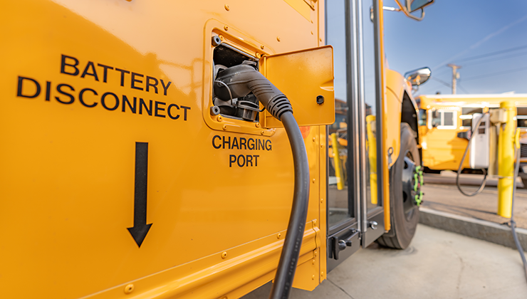 Side panel of a bright yellow school bus with electric charging nozzle plugged in and black lettering to the side "BATTERY DISCONNECT" with an arrow pointing down.
