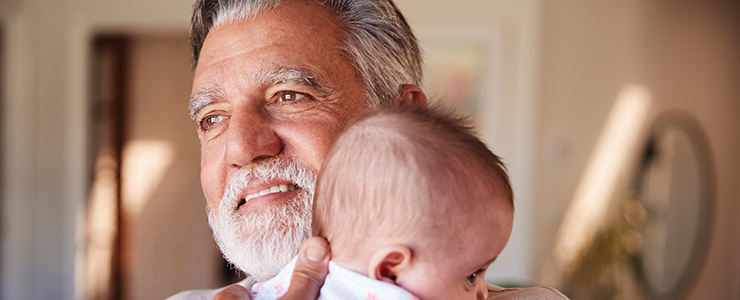 A smiling, white-bearded man gazes out on a sunny day, embracing an infant to his chest