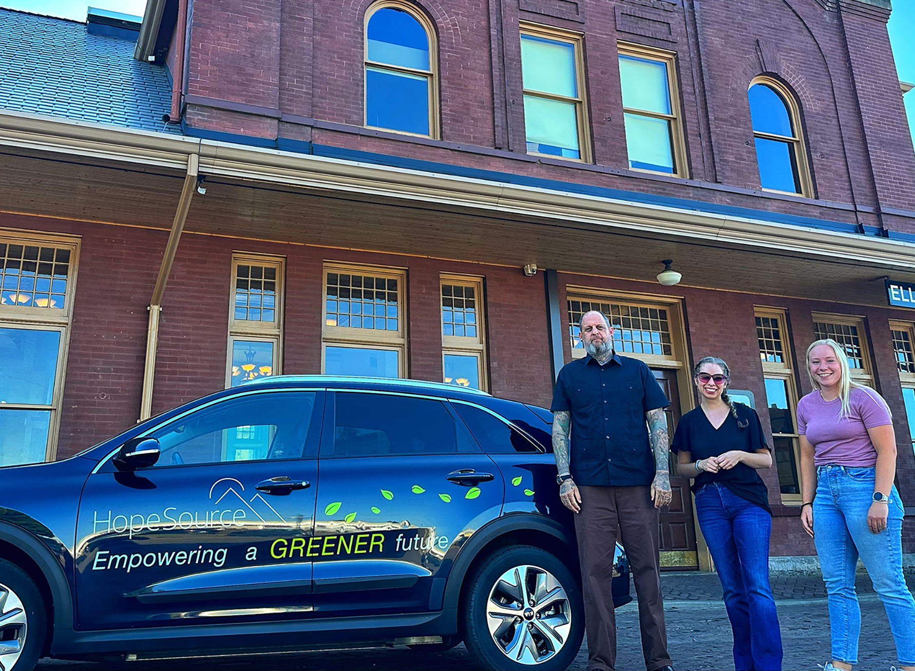 Two women and a man stand next to a "Hopesource" branded electric car, in front of a brick building.