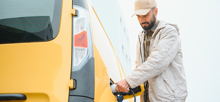 A man wearing a visored cap and windbreaker jacket holds an EV charging nozzle plugged into large yellow fleet vehicle.