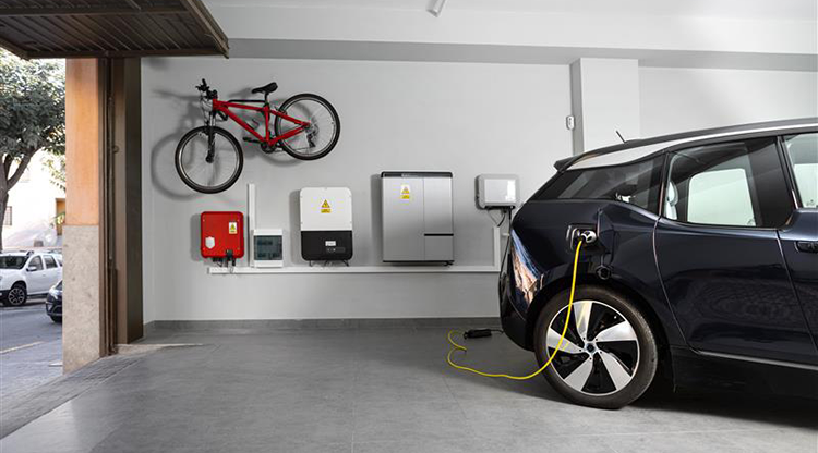 A black electric car is parked inside a home garage, charging cable attached, electric charging and storage panels line the wall, next to a red bicycle. Green tree lined neighborhood street is visible through the raised garage door.