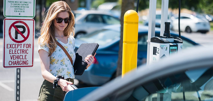 A woman wearing sunglasses and holding a black portfolio notebook, holds the nozzle of an EV charger plugged into a compact car in a large parking lot