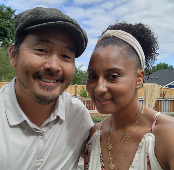 Nicole and her husband, smiling, with a blue sky and green neighborhood trees in the background