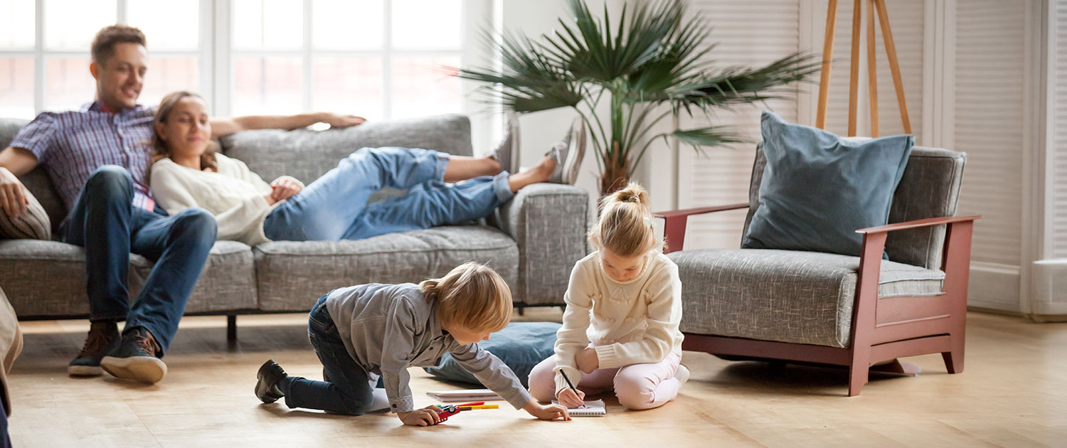 Boy and girl children play with crayons on floor and parents sit on sofa watching them