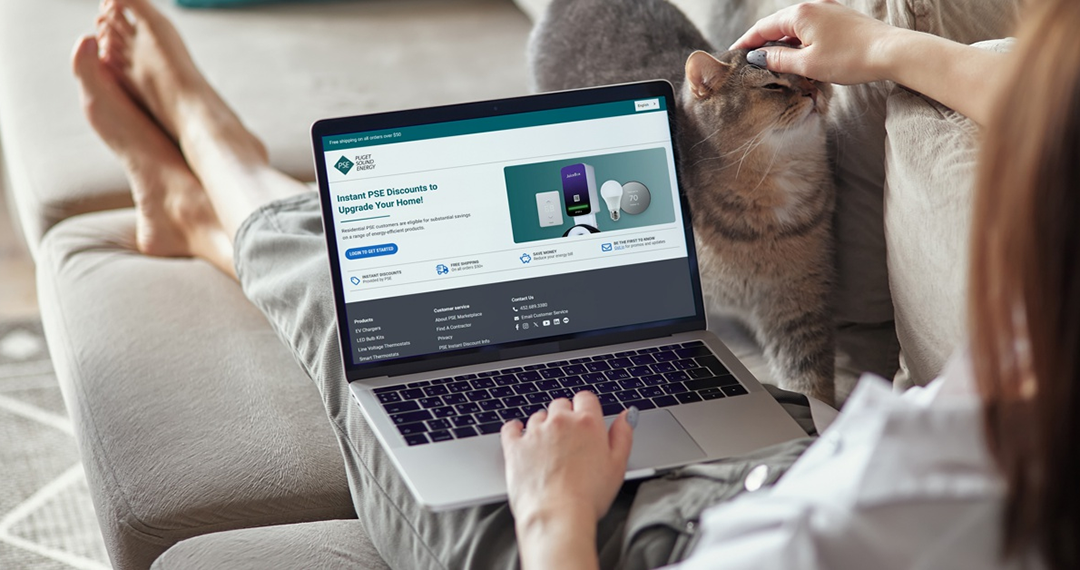 A barefoot customer relaxes on a couch with their cat while visiting the PSE Marketplace website on their laptop.