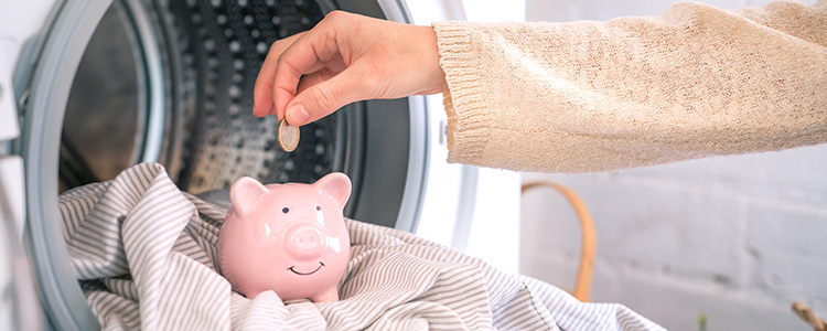 Hand drops coin into piggy bank sitting inside a dryer full of clothes